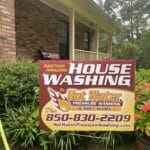 A yard sign advertising house washing services, placed in a garden in front of a home.